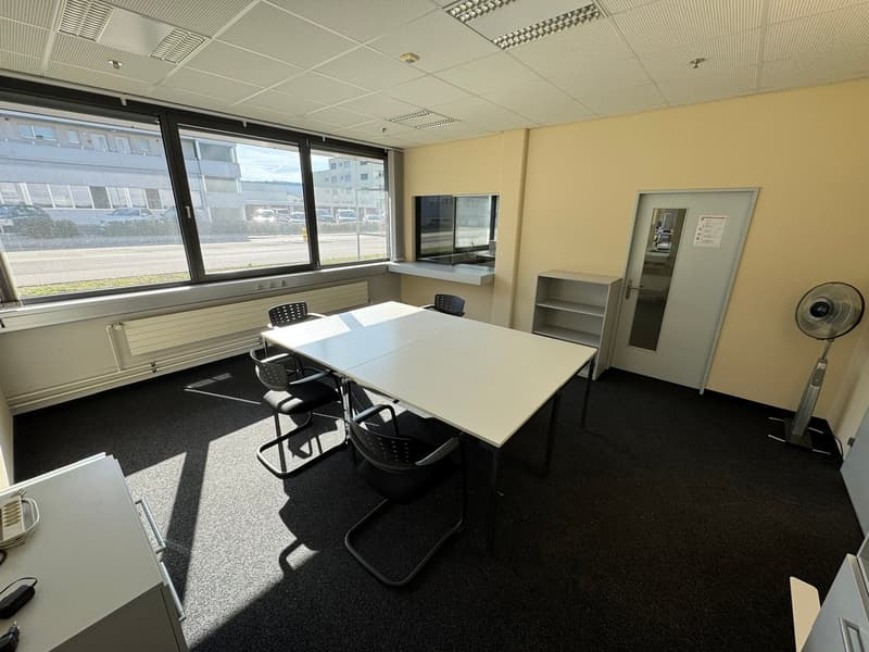 white walls, windows, gray carpet, white table, chairs, bookshelf, fan, white equipment on the left