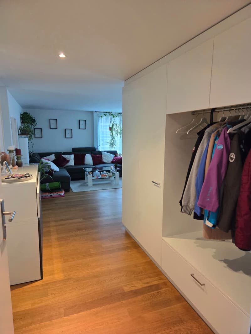 Entrance hall with wooden floor, white wardrobes, clothes hanging, living room with sectional sofa, coffee table, and plants