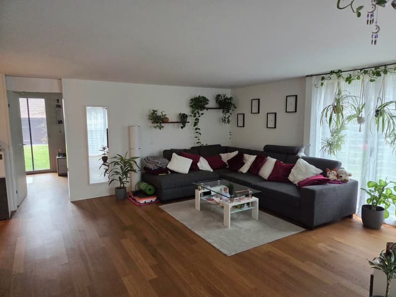 L-shaped sofa, glass coffee table, rug, wooden floor, white walls, plants, framed pictures, sliding door, glass door