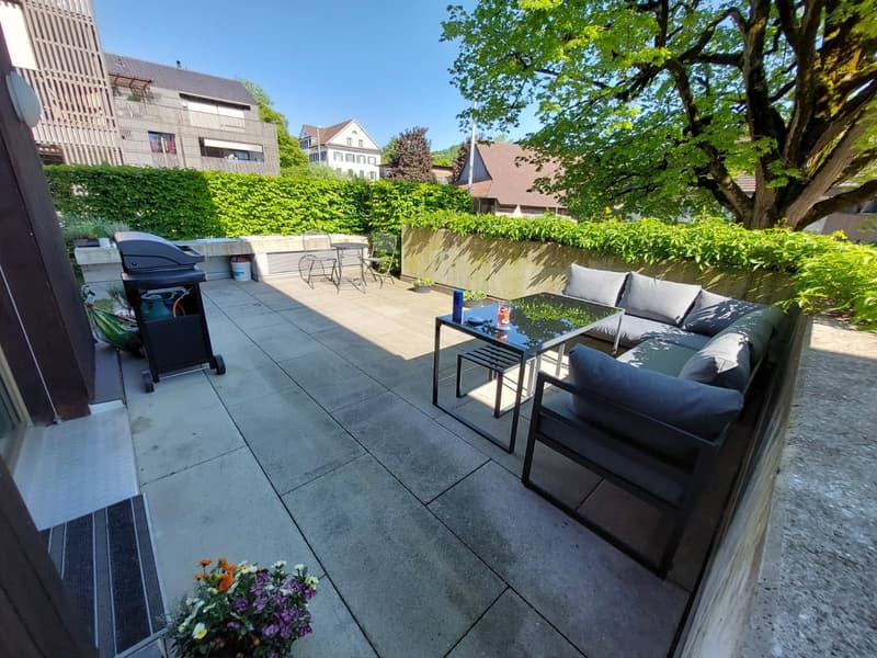 Outdoor patio with a sofa, table, and grill, surrounded by greenery and a tree