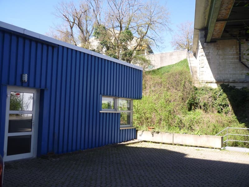 Blue building with a white door and a window, paved parking area in front, a slope with greenery behind