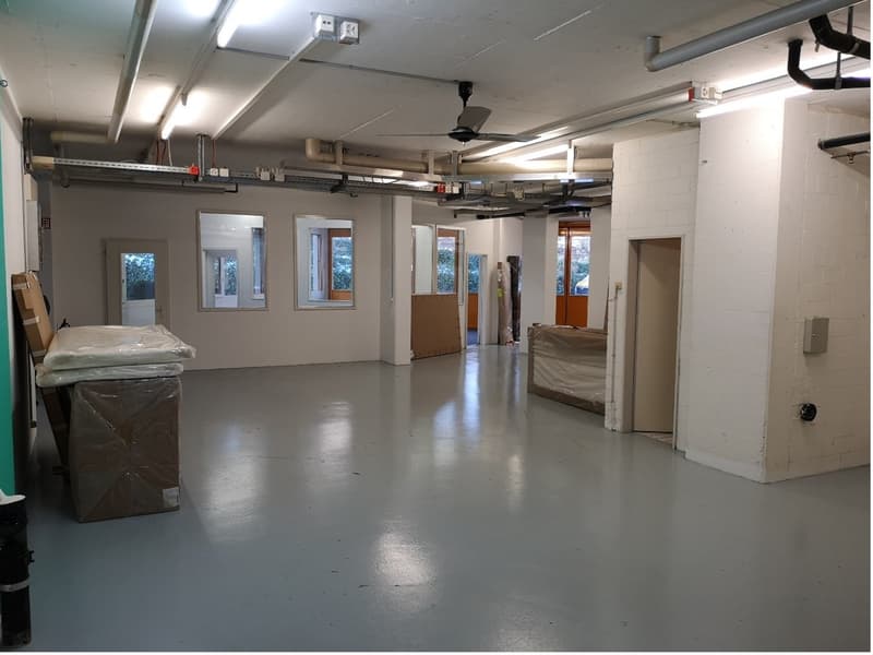 an empty room with gray floor, a ceiling fan, some windows, and doors, there are a lot of exposed pipes on the ceiling and walls, some objects and storage boxes on the side