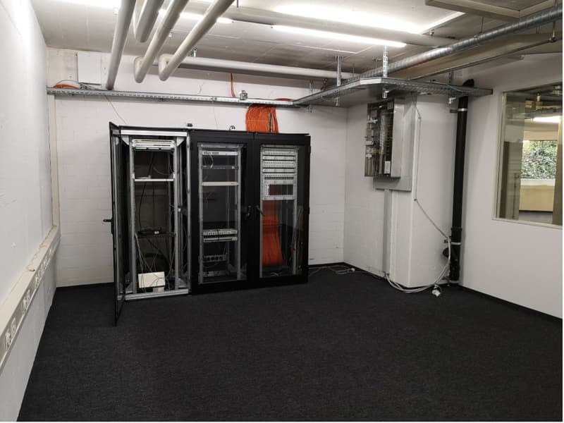 Server room with black racks, pipes and electrical wires, carpeted floor, windows on the right