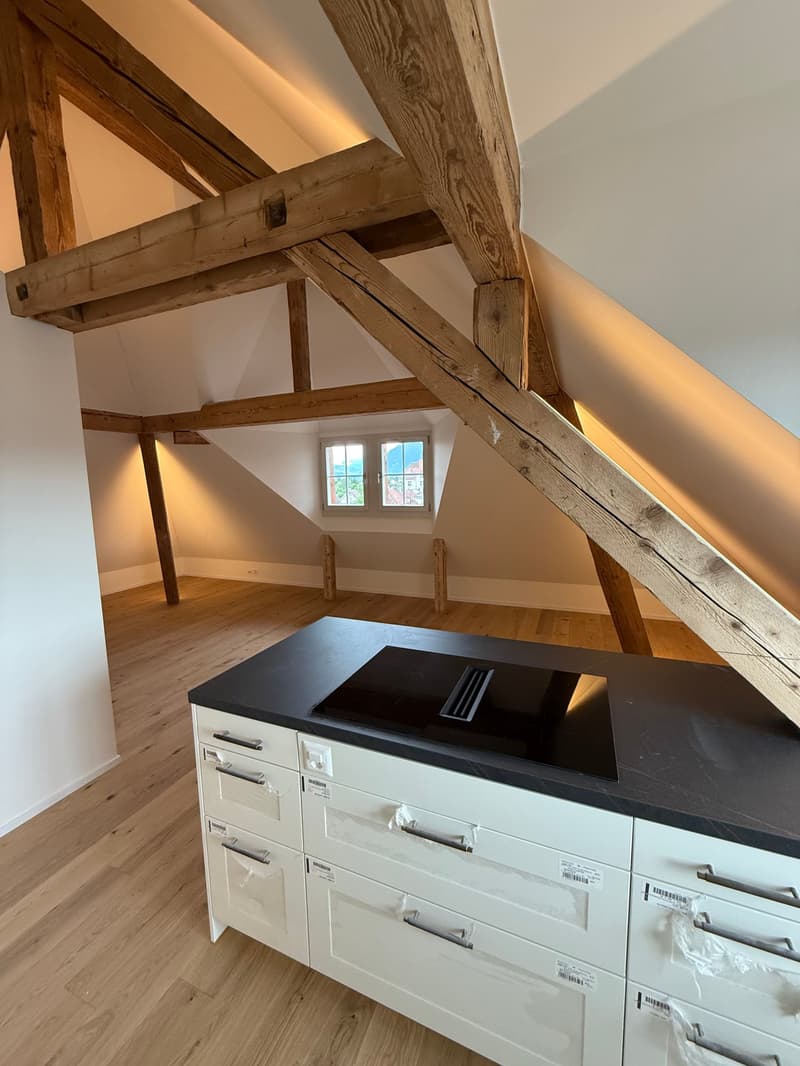 Attic space with exposed wooden beams, hardwood flooring, and a built-in cabinet with a black countertop. The room has a window providing natural light.