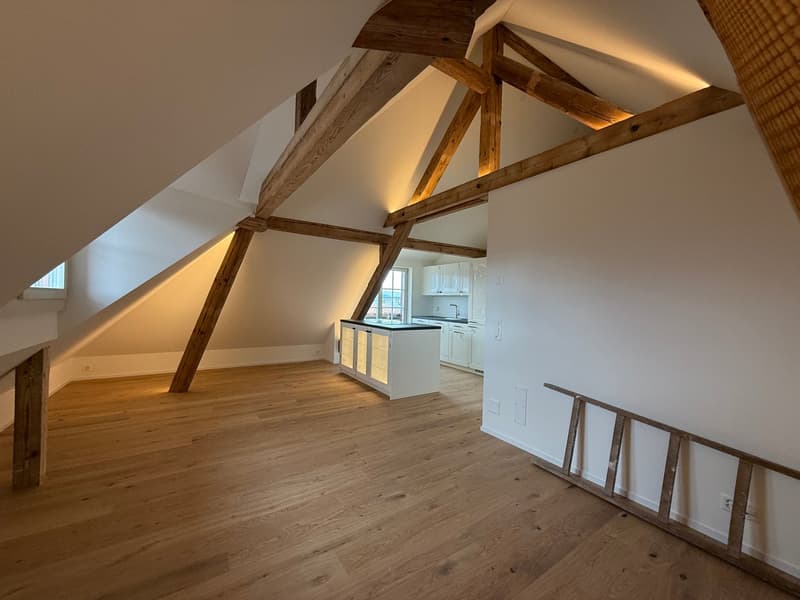 Spacious attic or loft space with exposed wooden beams and trusses, hardwood flooring, and a kitchen area with white cabinets visible in the background.
