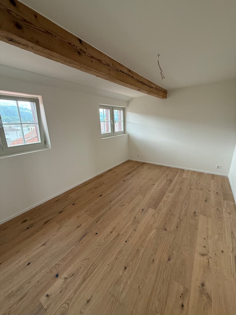 Large empty room with hardwood floor, white walls, large windows, wooden beam on ceiling