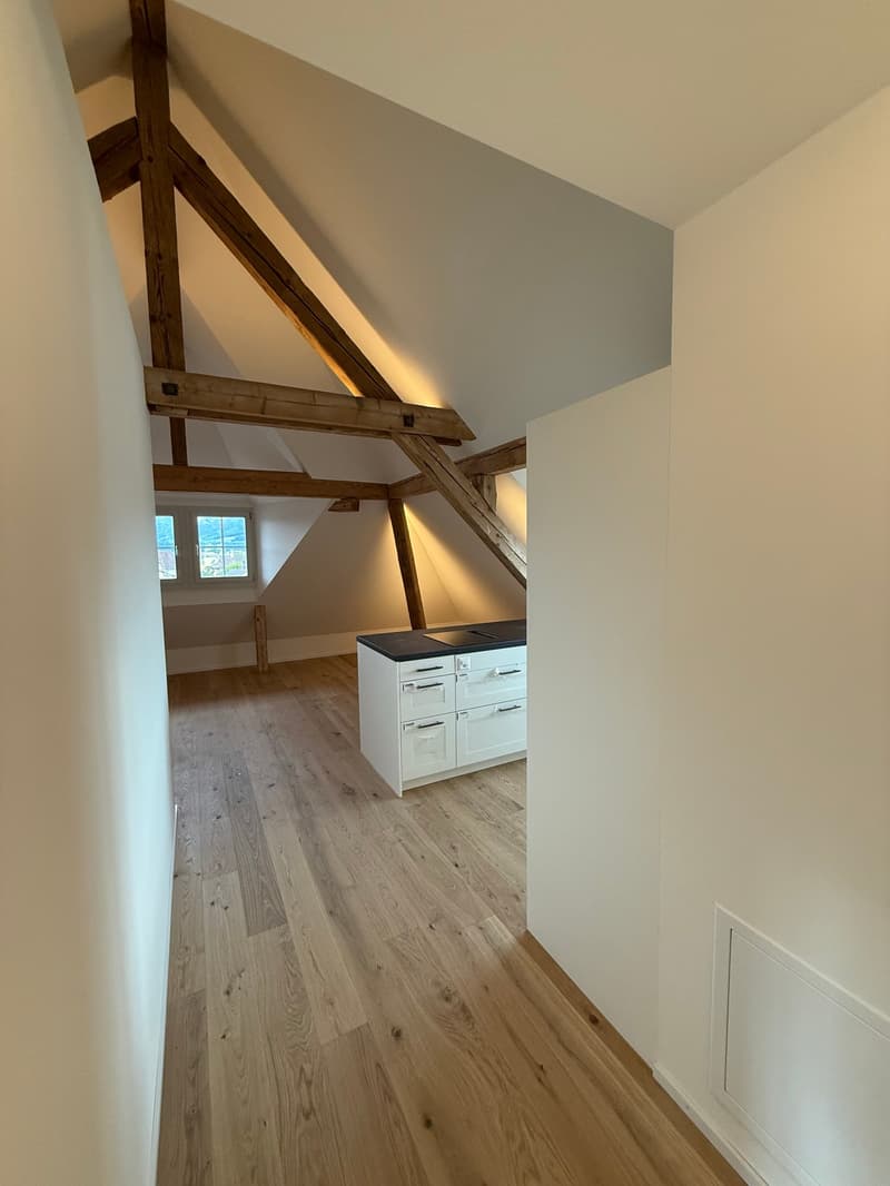 Attic space with exposed wooden beams, hardwood flooring, and a built-in cabinet or storage unit. The room has a cozy and rustic feel with the combination of the wooden elements and the gray walls.