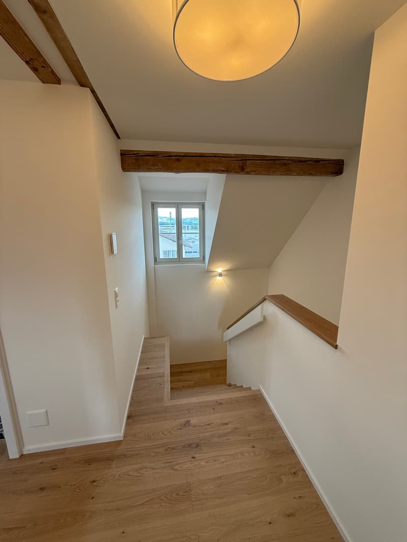 The image shows a staircase with wooden steps and a wooden beam running along the ceiling. The walls are painted white, and there is a circular light fixture hanging from the ceiling.