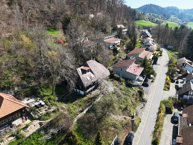 A hillside residential area with several detached houses of varying sizes, surrounded by trees and greenery. Each house is situated on a hill, with one appearing to be slightly raised compared to the others. The surrounding area is lush with green grass a