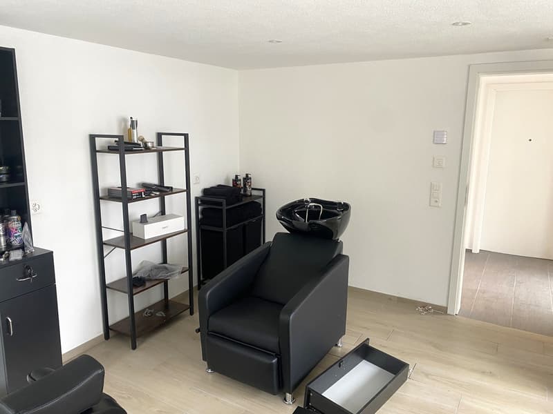 This image shows a room with a black salon chair, a black shelving unit, and a black cabinet. The room has white walls and a light-colored hardwood floor. There appears to be a private washing machine and a dishwasher visible in the room.