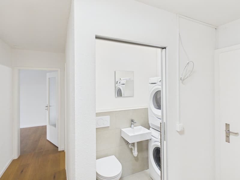 modern bathroom with toilet, white sink, wall-mounted washer, dryer, small mirror, white walls, wooden floor