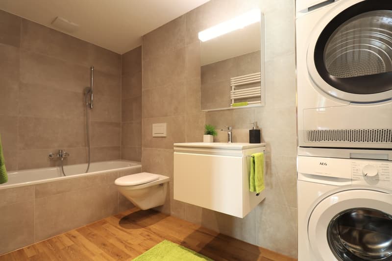modern bathroom with white fixtures, bathtub, sink, mirror, green rug, and stacked washer and dryer