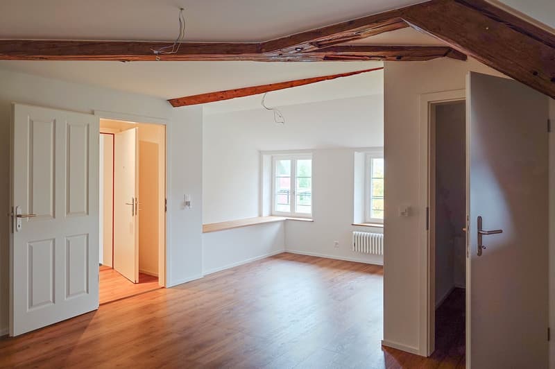 empty room, wooden floors, white walls, exposed wooden ceiling, two windows, wooden beams, radiators, wooden door frames