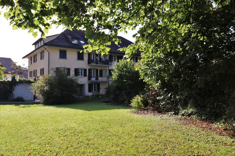 3-story house, yellow exterior, black roof, large balcony on the second floor, front garden with green grass and bushes