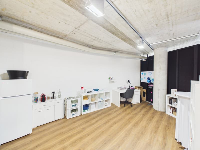 A room with white walls, wooden floors, and a large white fridge. Shelves and cabinets are against the wall. A desk with a black chair and a black lamp is placed next to the wall. There is a bucket, storage boxes, and bottles on a small table beside the f