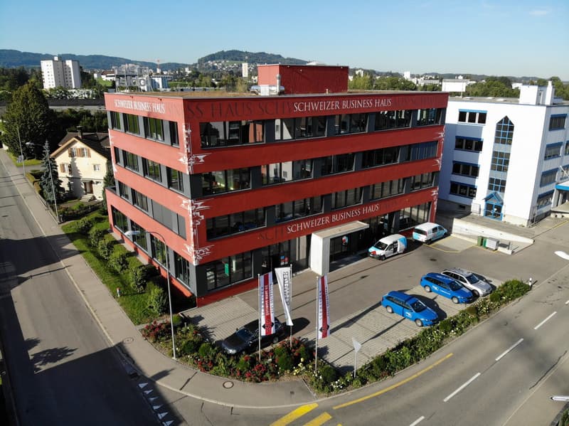 Red office building, multiple floors, multiple windows, parking lot with several cars, street light, greenery