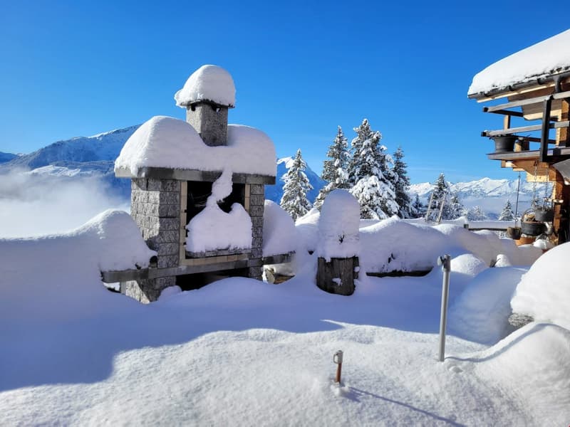 Wooden structure with stone base, covered in snow, snow-covered mountains in the background, blue sky