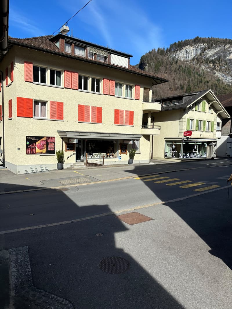 Multi-story building with yellow exterior walls, red shutters, and a balcony. There are shops and businesses on the ground floor, with residential units above. The building is situated in a mountainous area with a rocky cliff in the background.
