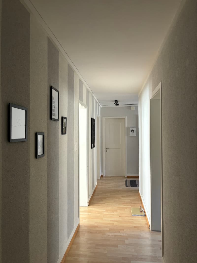 The image shows a long, narrow hallway with white walls and a hardwood floor. There are several framed pictures or artwork hung on the walls, and a doorway can be seen at the end of the hallway.