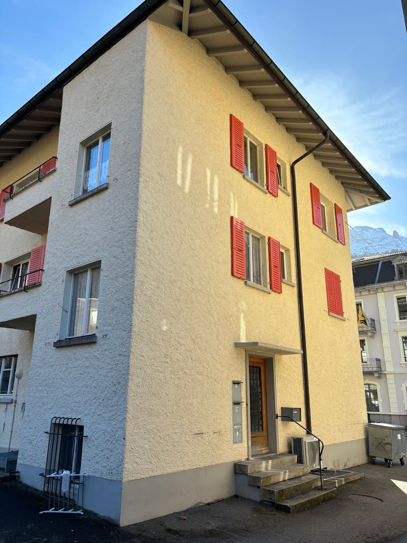 A multi-story residential building with a stucco exterior and red shutters on the windows. The building has a slanted roof and appears to be located in a mountainous area with a clear blue sky in the background.