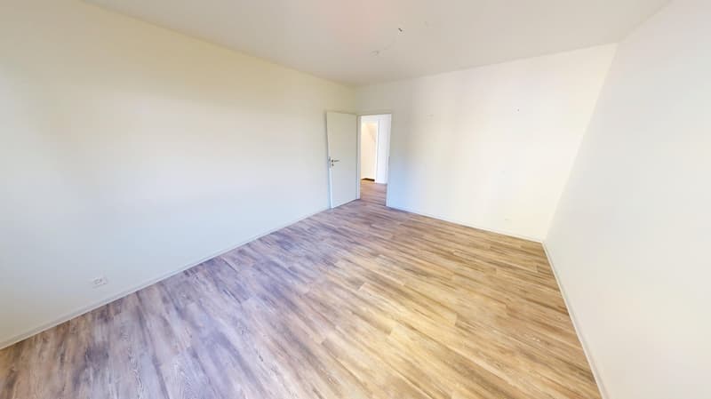 An empty room with wooden floors and white walls. A door leading to another room.
