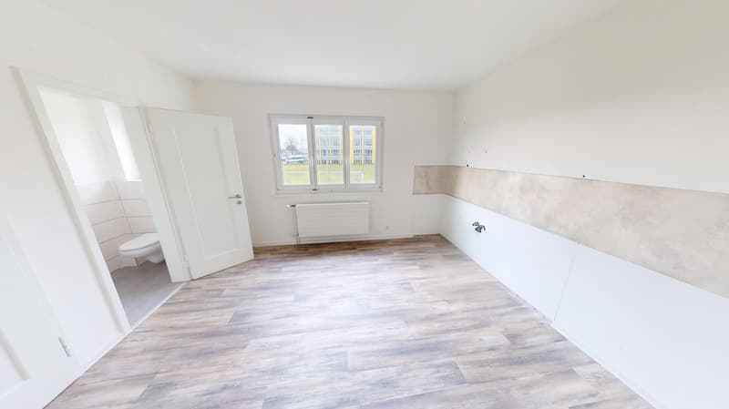 Empty room with wooden floor, white walls, radiator, open door, visible toilet in corner.
