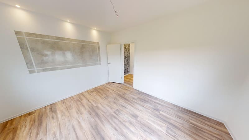 An empty room with wooden flooring, white walls, a white door leading to a hallway, and a wooden floor in the hallway.