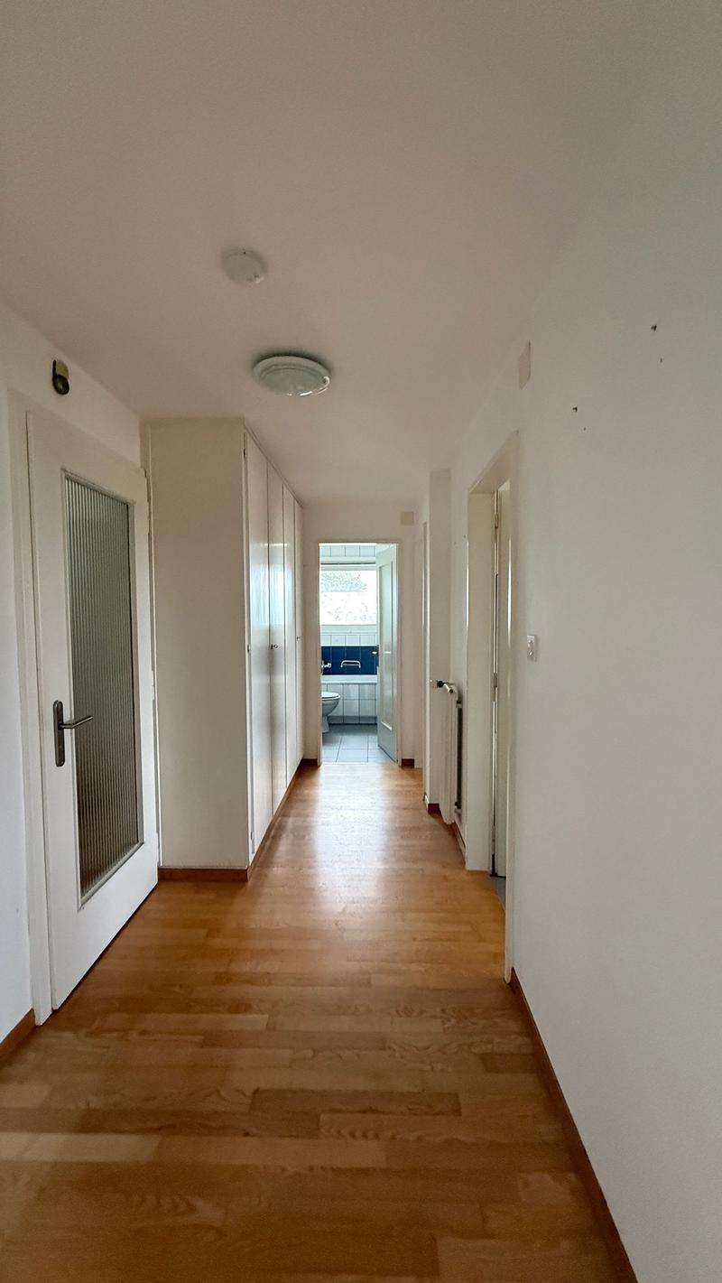 Empty hallway with white walls, wooden floor, closed doors, recessed lighting, bathroom glimpse.