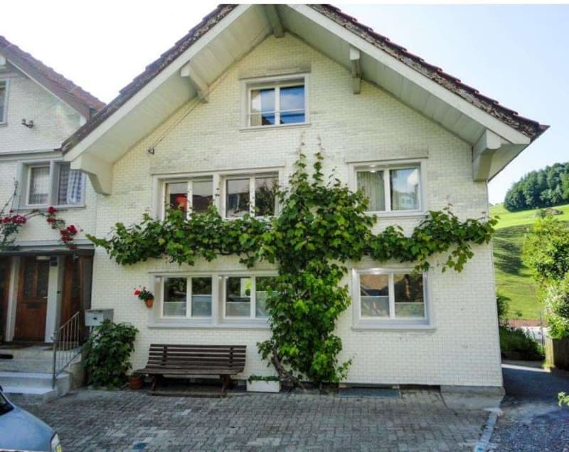A white, two-story house with a brown roof and white windows covered by climbing plants. There is a small garden with a wooden bench and plants to the right of the house. The front has brick-paved driveway and a parked car. The house has steps leading to