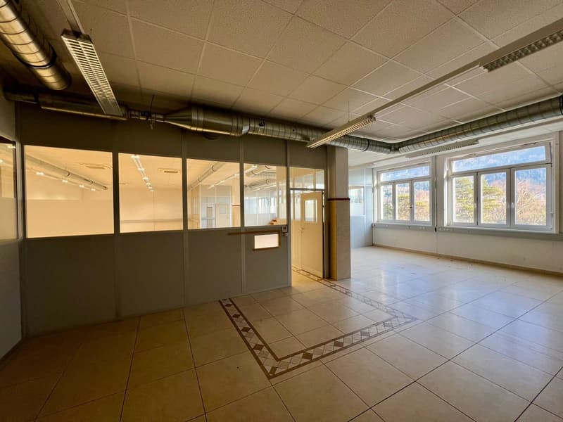 The image shows a large, open office space with a tiled floor, glass partitions, and large windows overlooking the outdoors. The ceiling has exposed ductwork and lighting fixtures. The space appears to be in a commercial or office building setting.