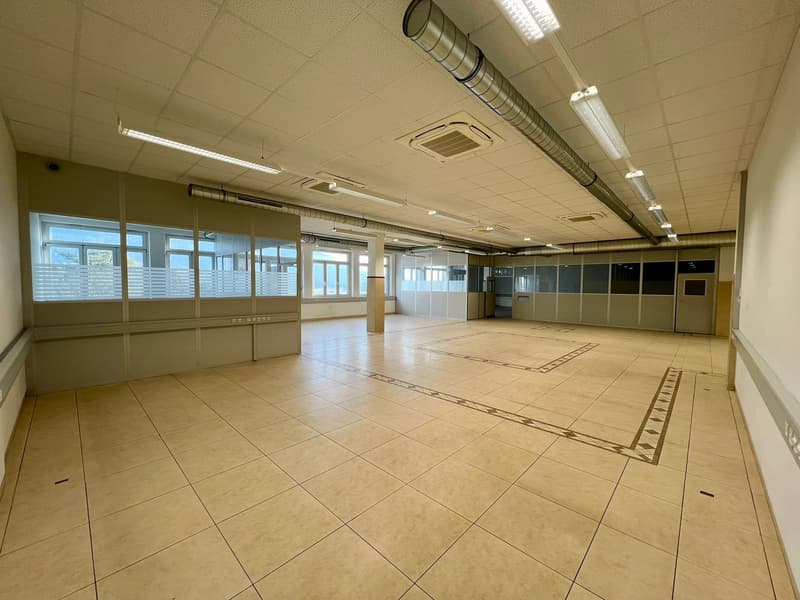 Large open office space with tile flooring, suspended ceiling with lighting, and glass partitions along the walls.