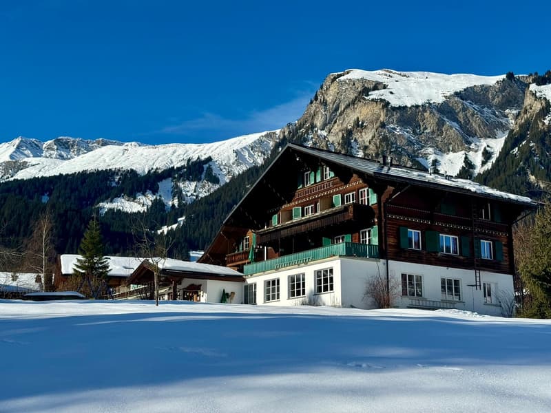 Wooden house, multiple windows, snow-covered ground, large mountains in the background