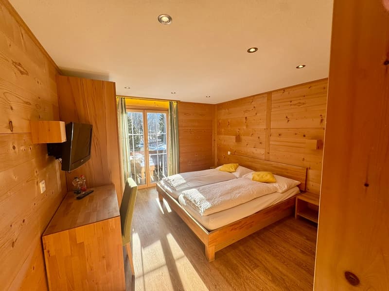 Bedroom with double bed, wooden walls, wooden floor, desk, mounted TV, small chair, balcony with glass doors, window with curtains, power outlet.