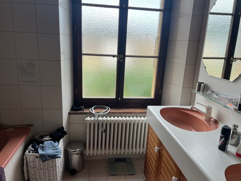 Bathroom with a sink, radiator, and a window with frosted glass. There is a laundry basket and some other items in the room.