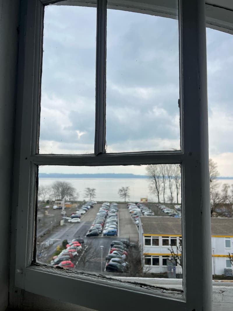 View of a parking lot with many cars parked outside through a window with a cloudy sky in the background