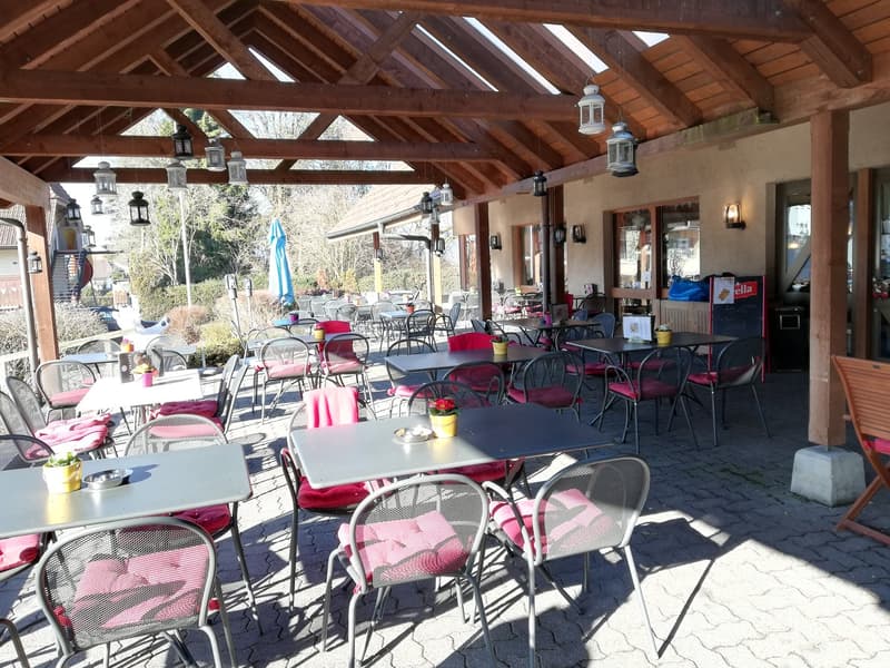 Covered outdoor dining area with wooden beamed ceiling, lanterns, and tables and chairs with red cushions. The area has a view of the surrounding nature through the open sides.