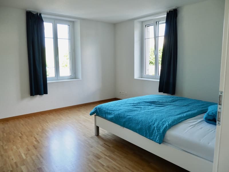 a room with a wooden floor, two windows with black curtains, a white bed with a blue bedspread and pillow