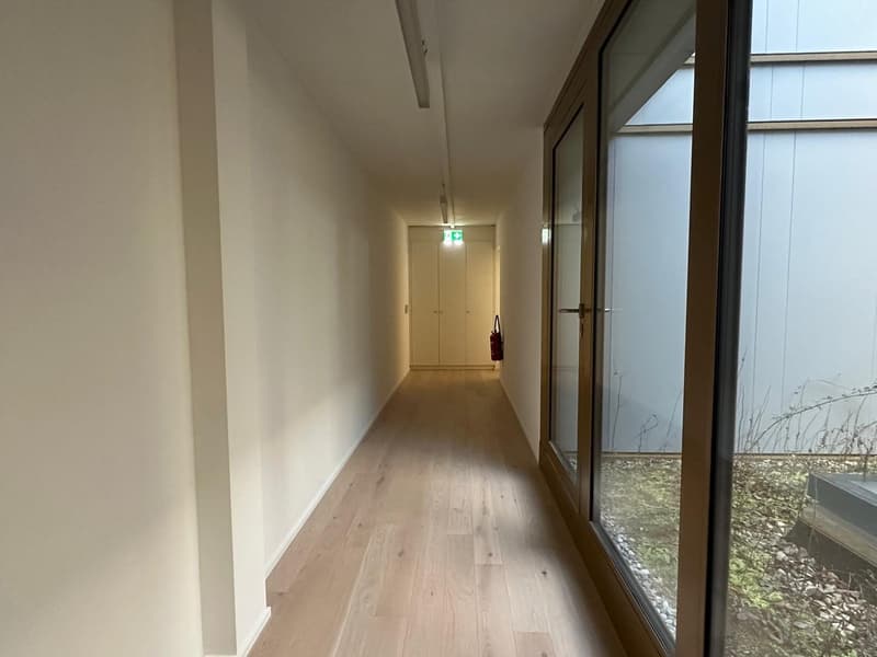 A corridor with wooden floors, white walls, an open door with a view to a garden, and a glass wall