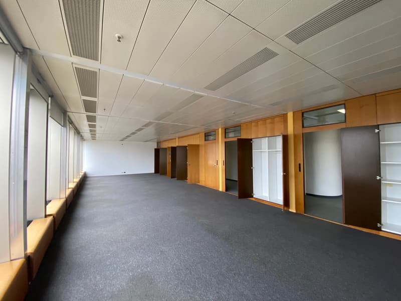 The image shows a long, empty office space with a gray carpeted floor and a suspended ceiling with recessed lighting. The walls are lined with wooden cabinets and doors, suggesting this is a commercial or office building interior.