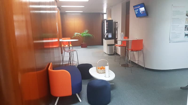 The image shows an office or lounge area with modern, colorful furniture. There are orange and blue chairs, a white table with a basket on it, and a vending machine or small refrigerator in the background. The walls are gray and there is a TV screen mount