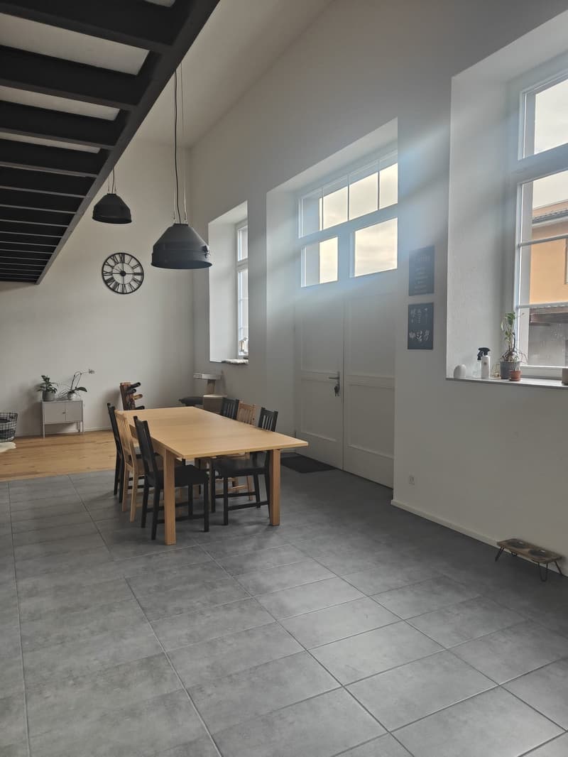 Dining room with wooden table and chairs, white walls, large windows, tiled floor, clock on wall, small cabinet with plant, black rug, black lamps