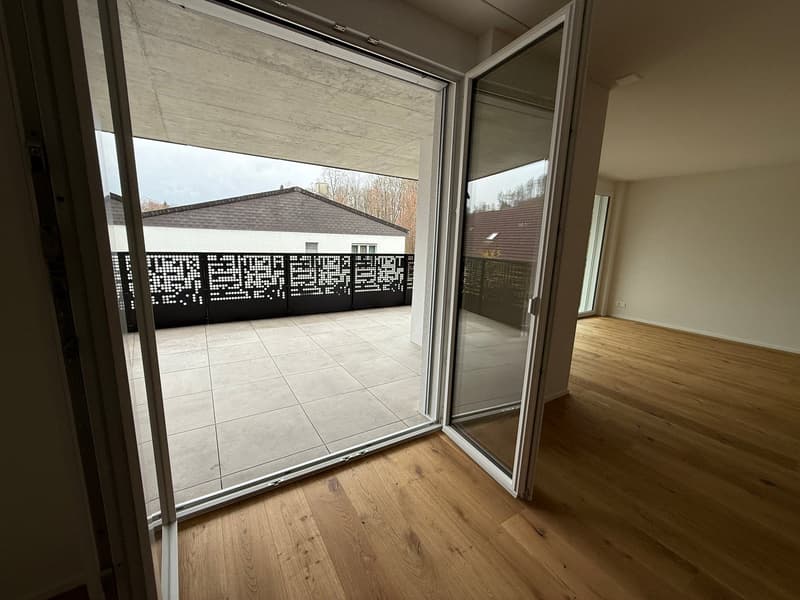 Open sliding glass doors leading to a balcony, wooden floor, white walls, black railings on balcony