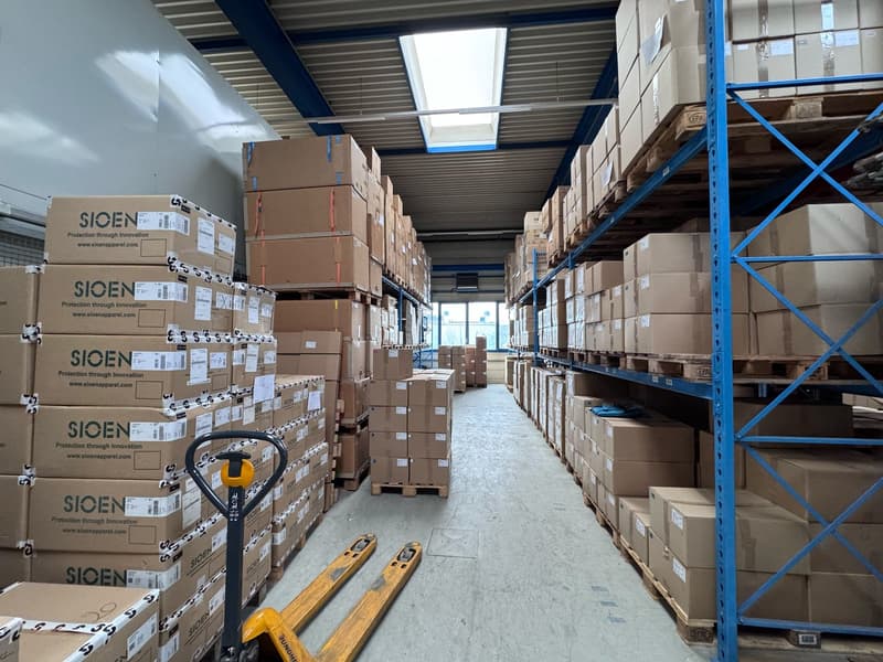 Large warehouse filled with numerous boxes, labeled 'SIONEN', and metal shelving units; a forklift is visible in the foreground.