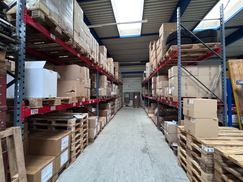 Warehouse with stacked storage boxes on either side, ceiling with skylights, and a pathway in the middle. Forklifts might be parked on the left side.