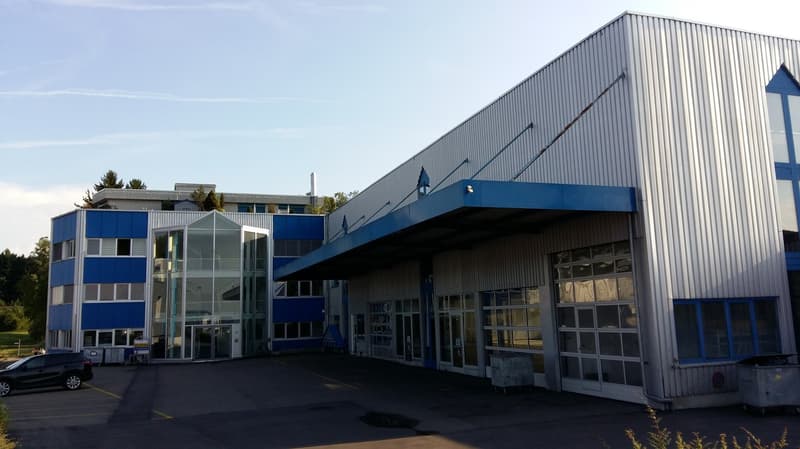 Large industrial building with gray exterior, blue and white trim, glass walls, multiple entry doors, large parking area.