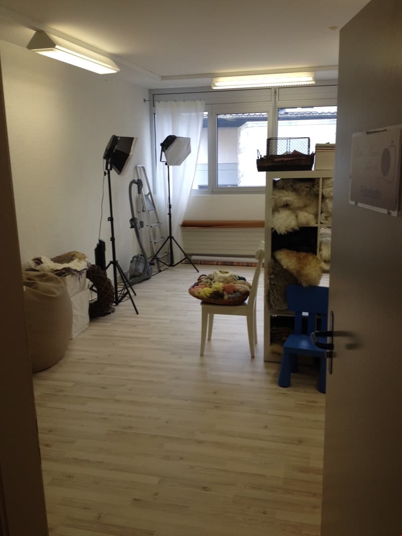 A room with wooden floors, white walls, ceiling lights, a window with white curtains, a storage box, a ladder, and a chair with a pillow on a basket.