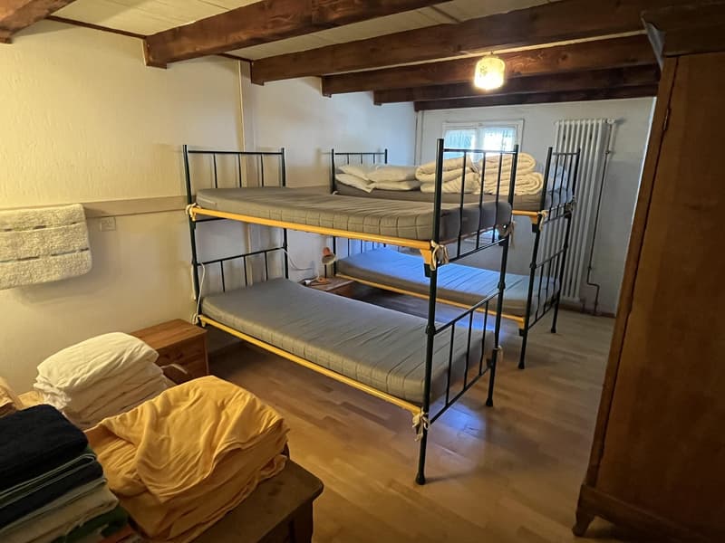 room with three bunk beds, mattress with white sheets, wooden floor, night table with lamp, and folded clothes on a table