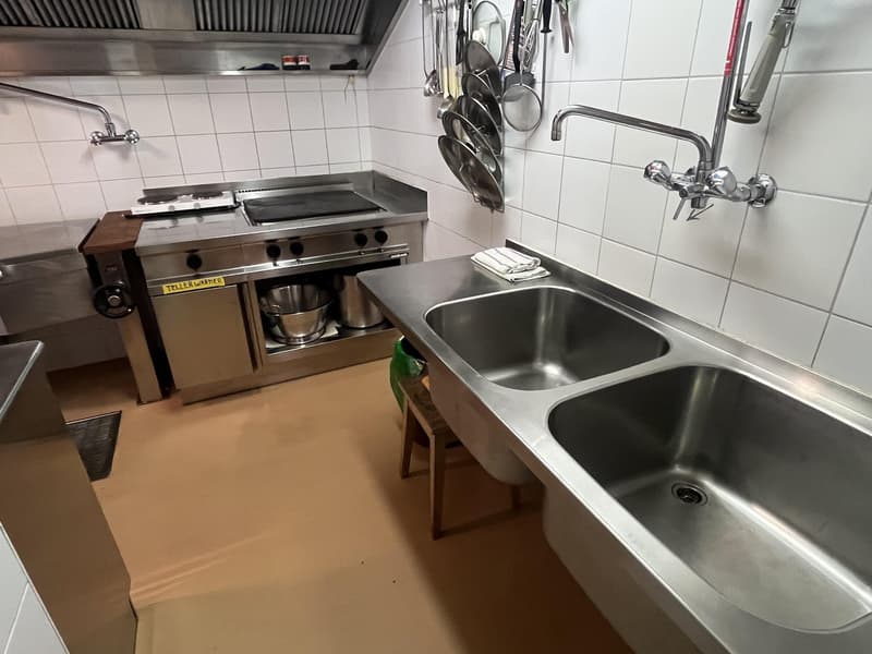 stainless steel sink, gas stove, two ovens, stools, pots hanging from the ceiling, tile walls, exhaust hood
