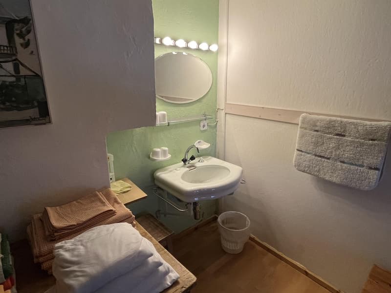 A small bathroom with a mirror, sink, towels, and trash bin. The wall is painted green, and there is a picture frame hanging on the wall. The floor is wooden, and there is a wooden bench. The sink is white, and there is a faucet. There is a mirror mounted