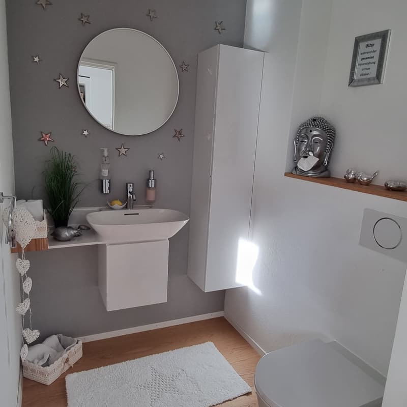 Compact bathroom with white sink, mirror, white cabinet, toilet, white floor mat, silver buddha figurine, and plants.